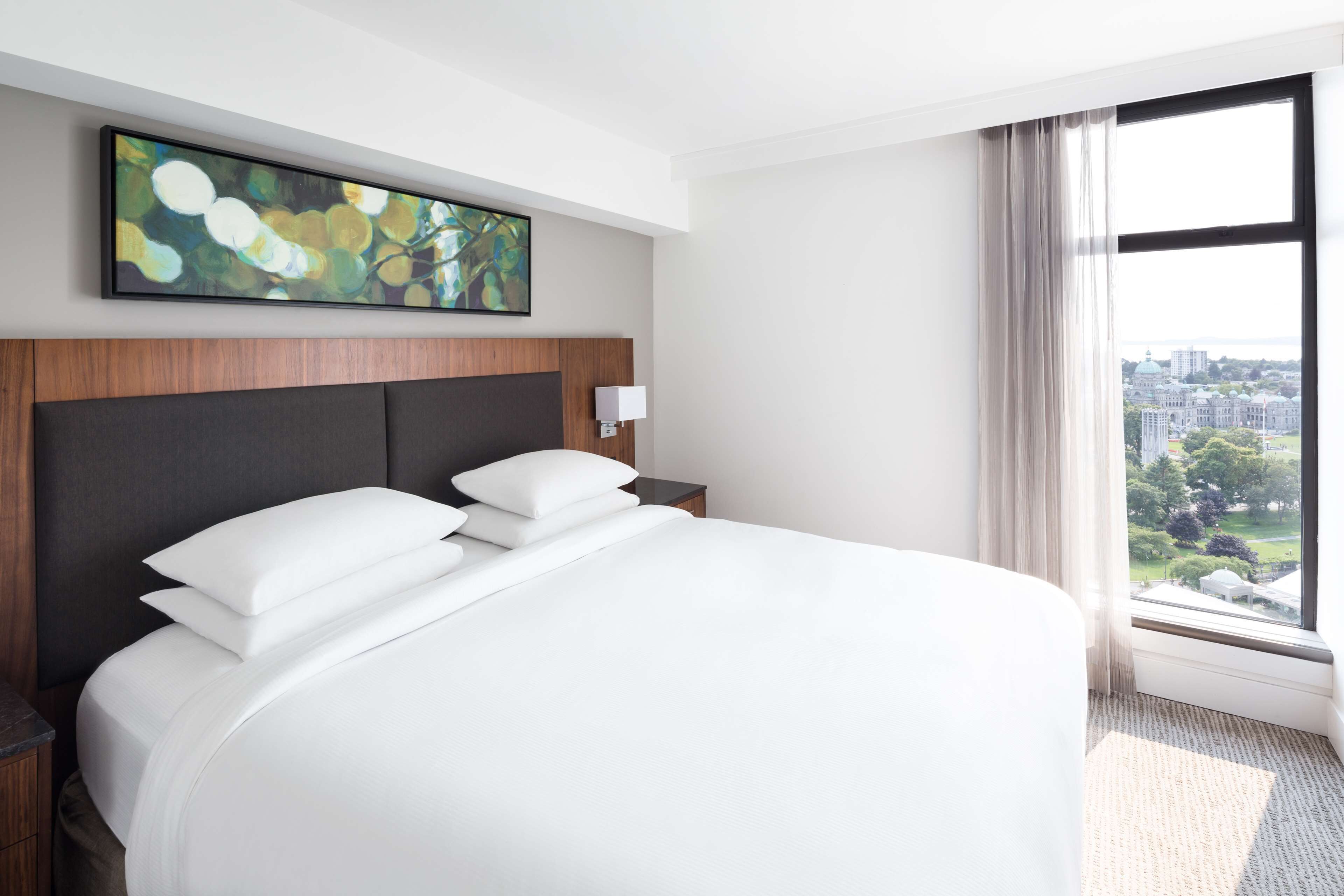 A hotel room with a large bed, a nightstand, a wall-mounted lamp, and a window with a city view.