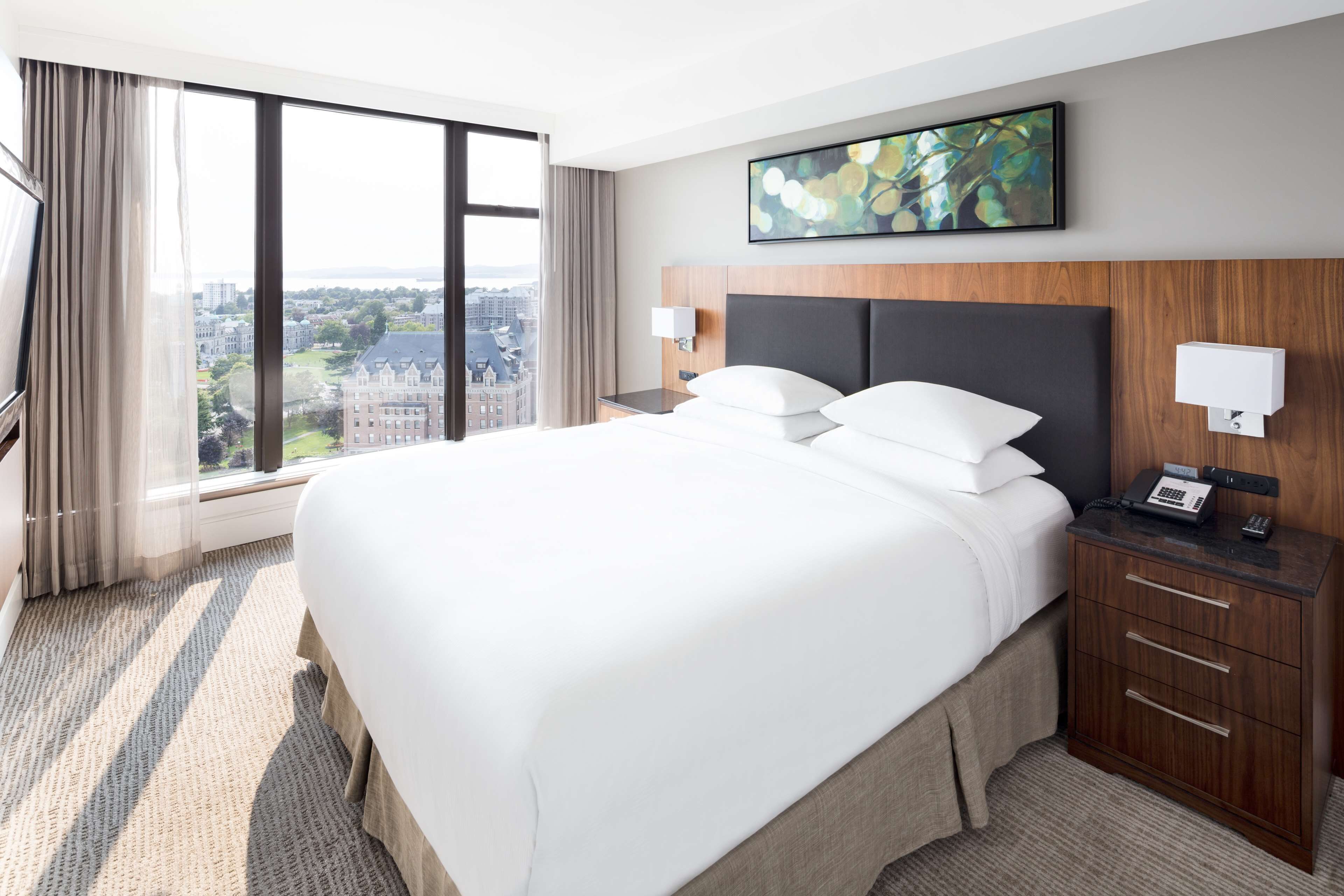 Habitación de hotel con una cama grande, una mesita de noche con lámpara, cabecera de madera y vista a la ciudad.