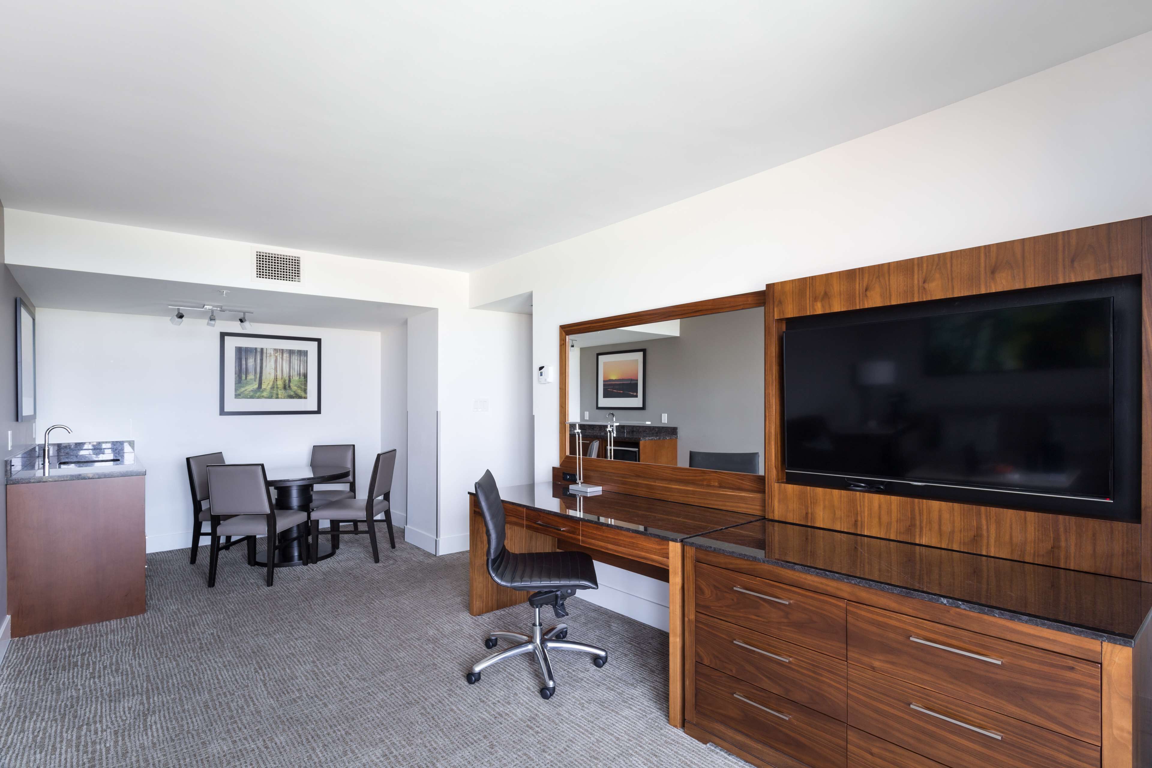 Ein modernes Hotelzimmer mit Schreibtisch aus Holz, einem Flachbildfernseher, einer Essecke mit Tisch und Stühlen sowie einer kleinen Küchenzeile.