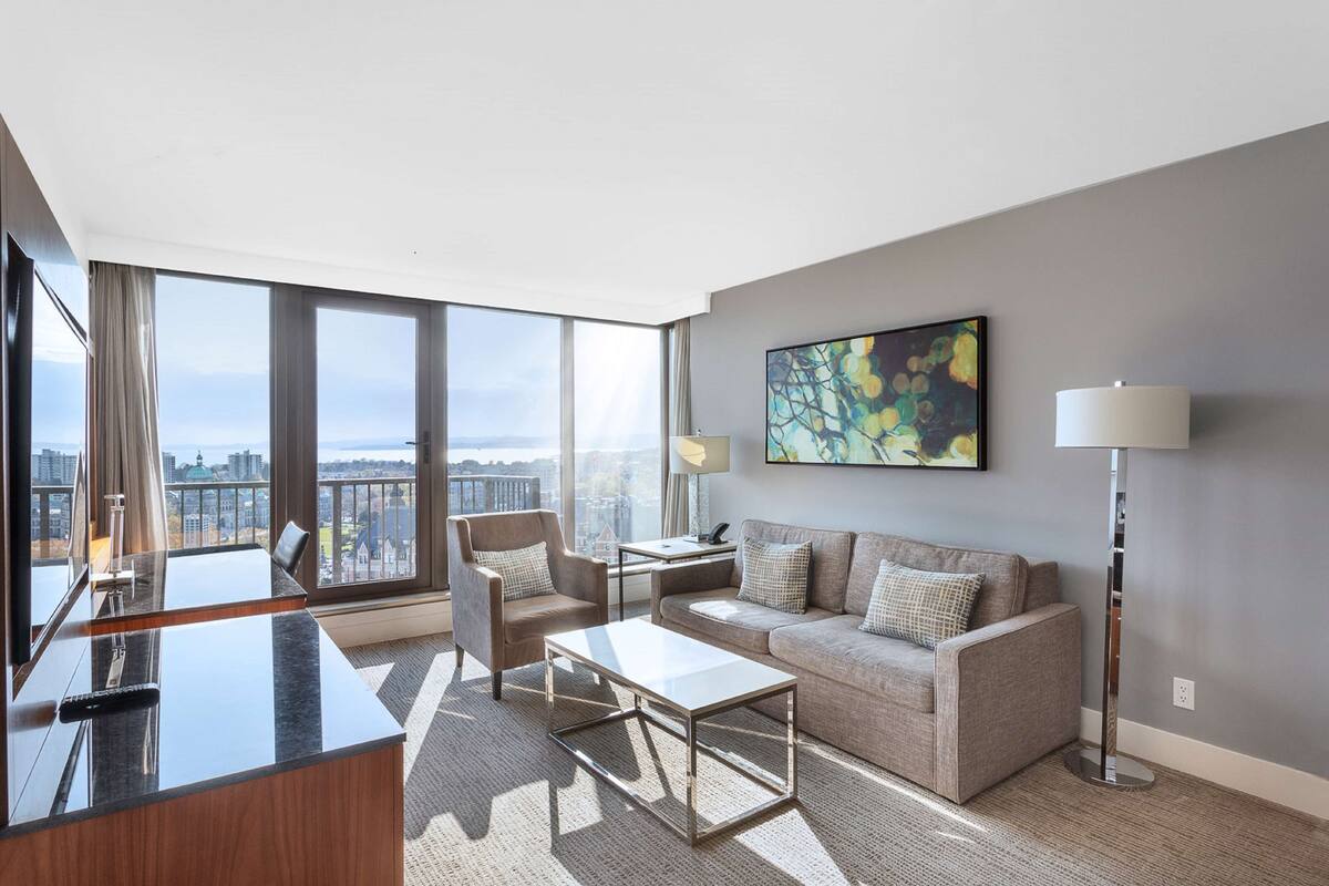 Una habitación de hotel moderna con un ventanal grande, un sofá, una mesa de centro, un escritorio y una lámpara.