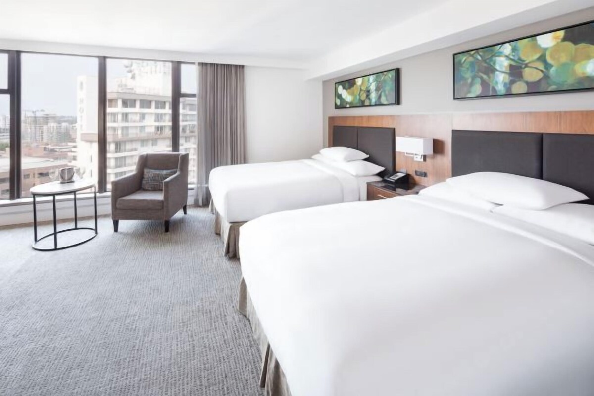 A hotel room with two beds, a chair, a small table, and a large window offering a city view.