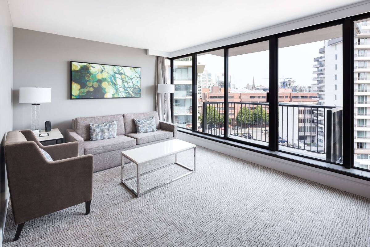 Una sala de estar moderna con sofá, sillón y mesa de centro, ventanas amplias con vista a la ciudad y un cuadro en la pared.