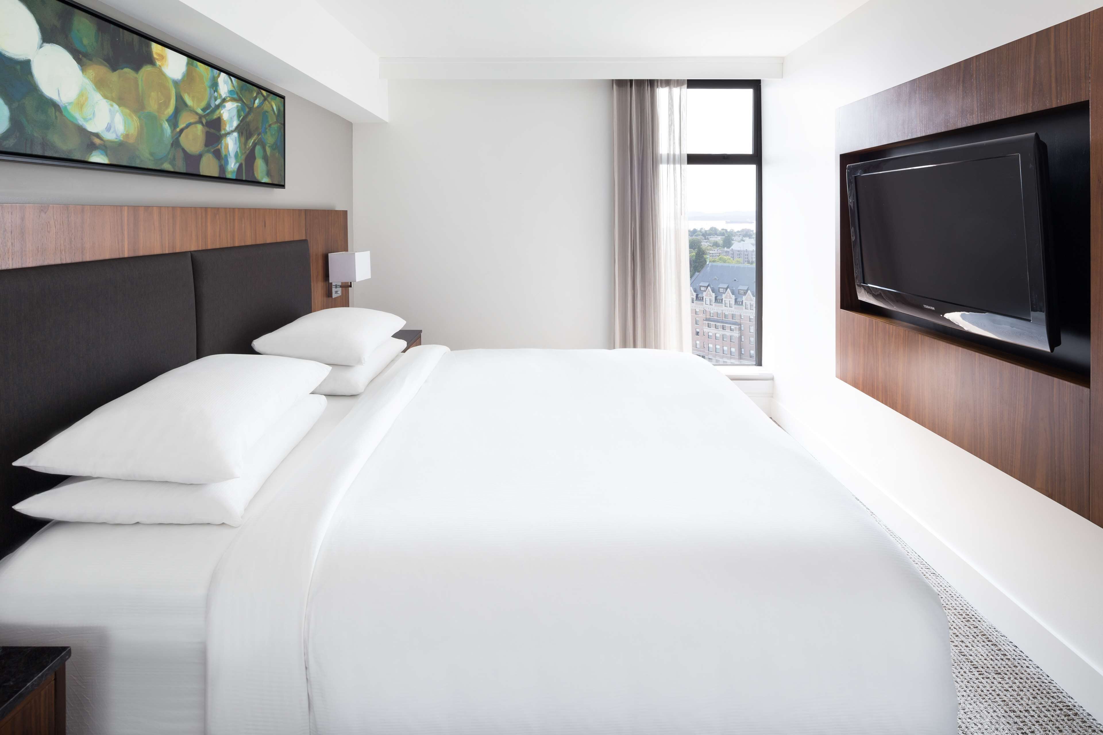Une chambre d’hôtel avec un grand lit, un téléviseur fixé au mur, un tableau au mur et une fenêtre donnant sur des immeubles.