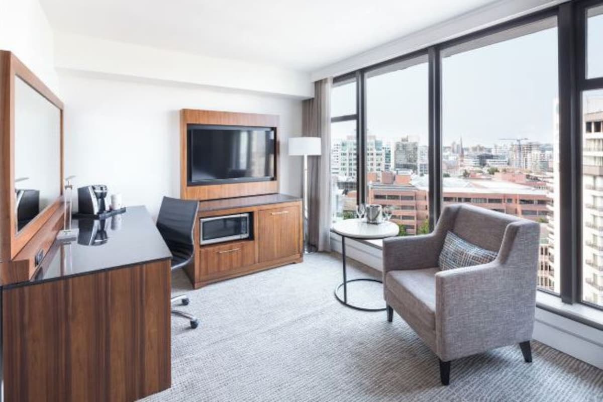 Quarto de hotel com escrivaninha de madeira, televisão de tela plana, sofá e vista para a cidade através de amplas janelas.