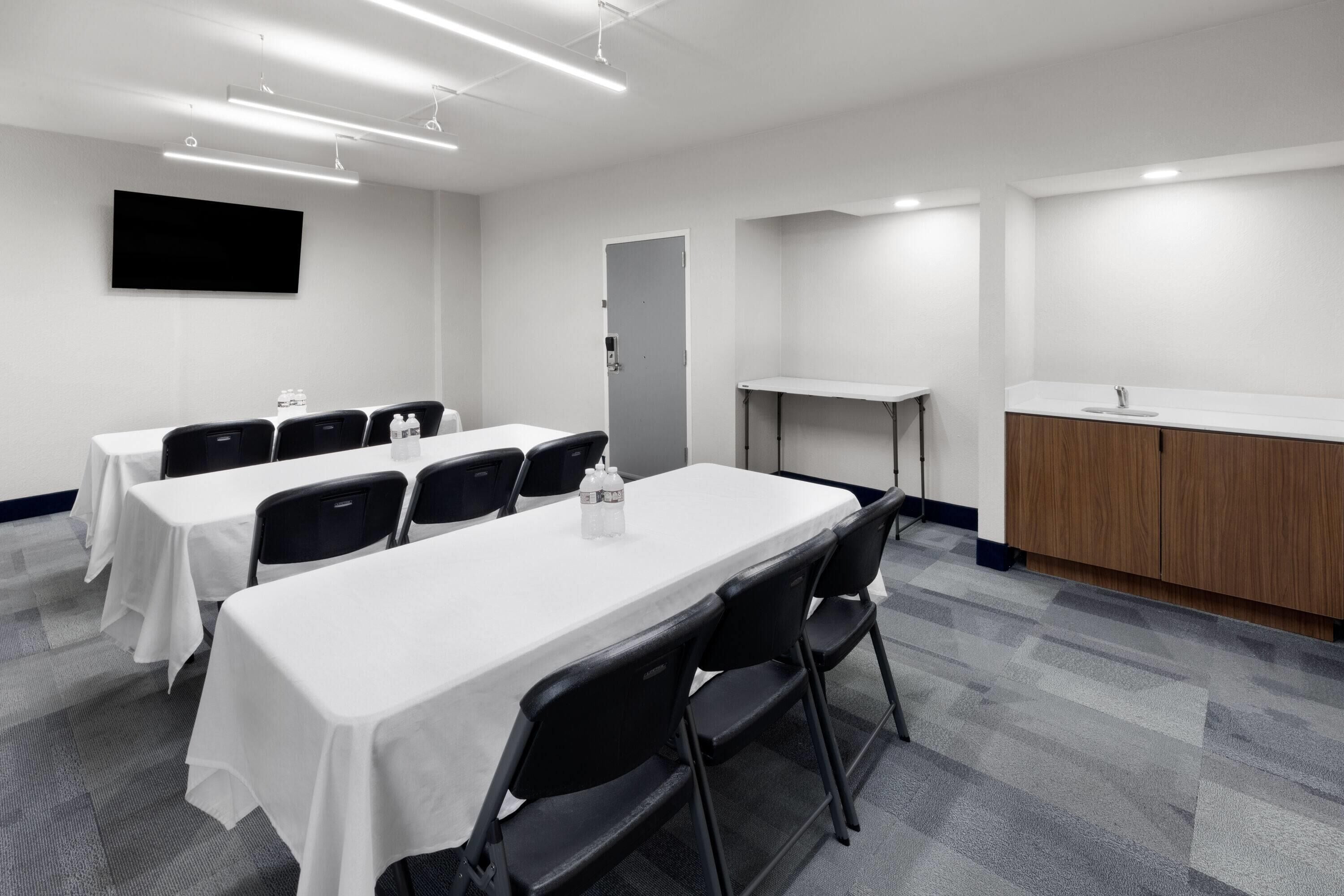 A conference room with a long table, chairs, a TV, and a kitchenette area.
