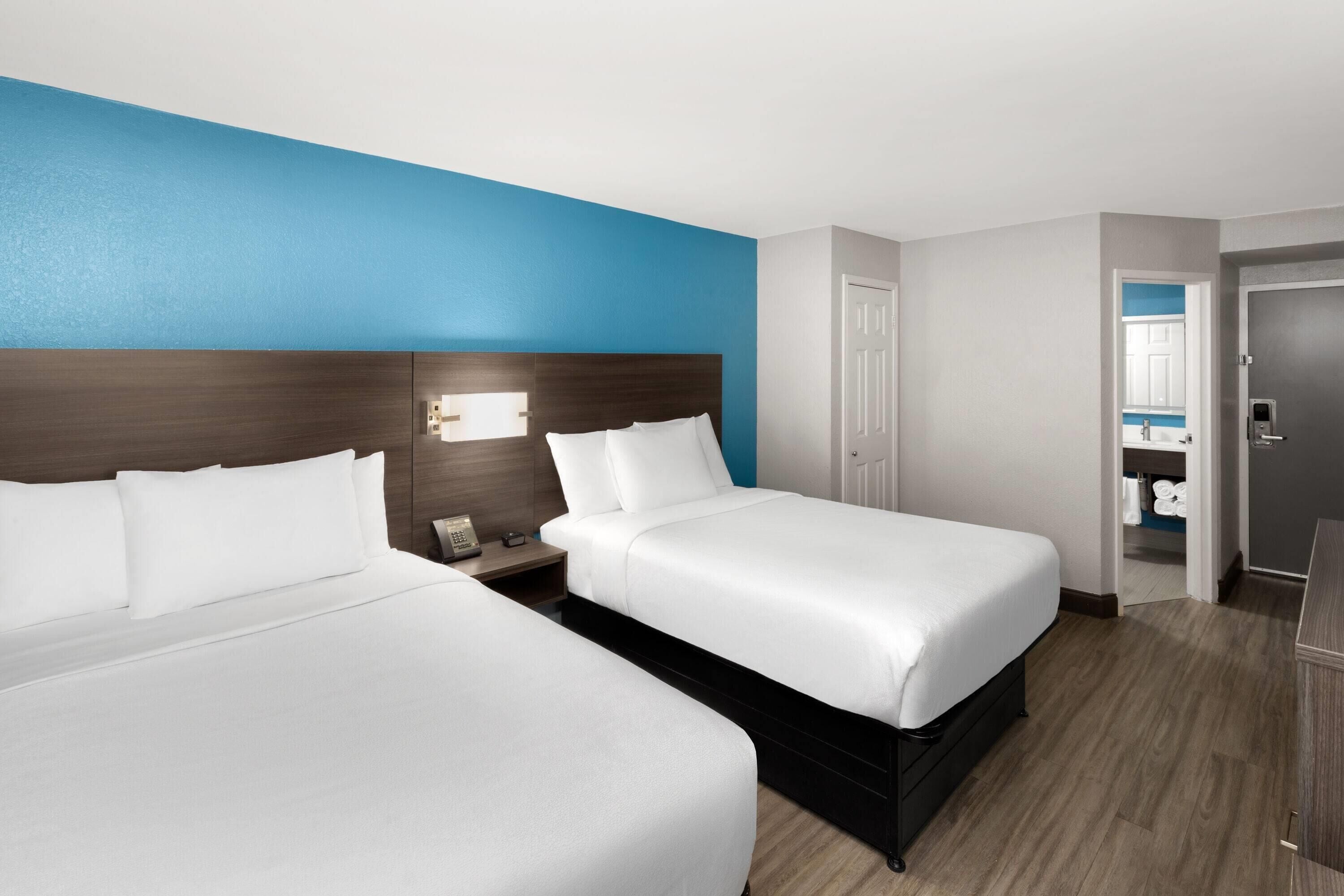 A hotel room with two beds, a blue accent wall, a wooden headboard, a nightstand with a phone, and a door leading to a bathroom.