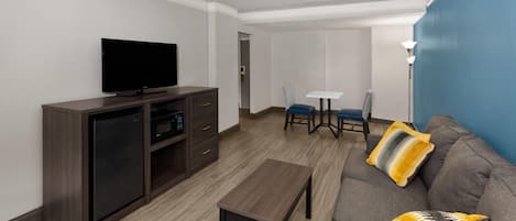 A hotel room with a flat-screen TV, a small refrigerator, a wooden coffee table, a gray sofa with yellow cushions, a small table with two chairs, and a blue accent wall.