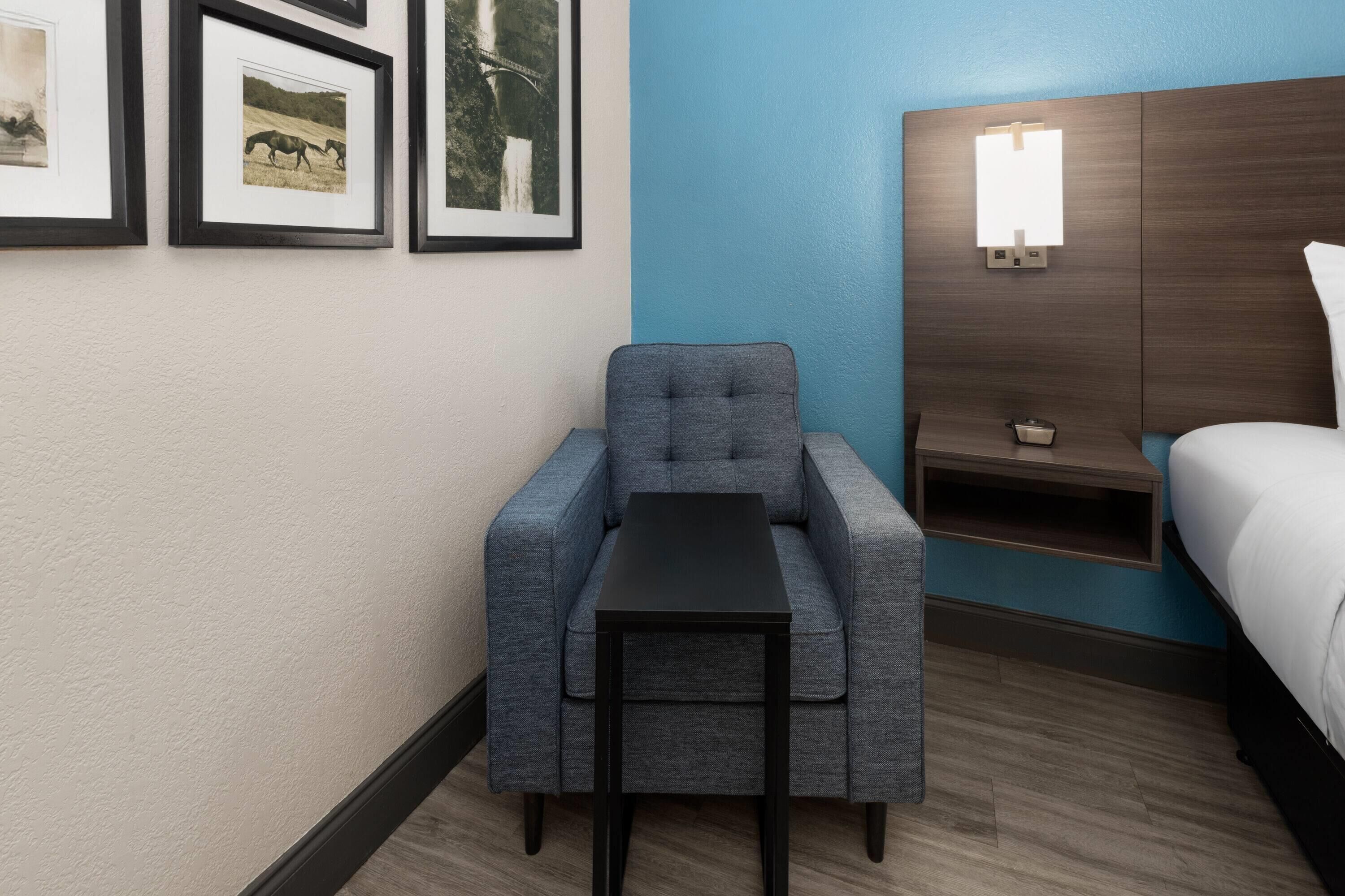 A hotel room with a grey armchair, a small black table, a bed with white bedding, a nightstand, and a wall-mounted light fixture.