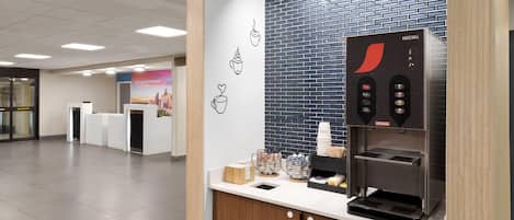 A modern coffee station with a Nescafé branded espresso machine, a counter with coffee cups and a basket of napkins, and a tiled wall with a cityscape poster.
