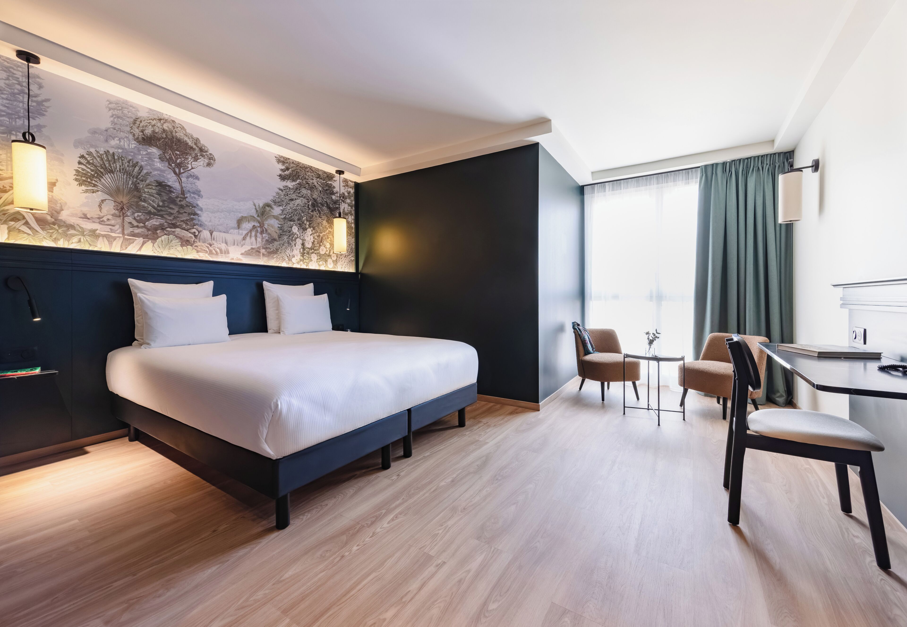 A hotel room with a bed, a desk, two chairs, a large window with curtains, and a wall mural.