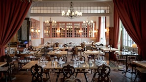 A restaurant with multiple tables and chairs, a chandelier, and a view of the kitchen in the background.
