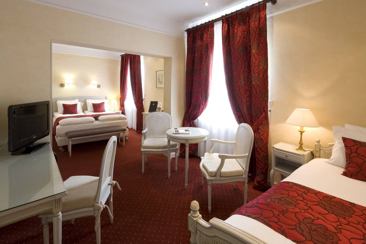 A hotel room with a bed, a desk with a television, two chairs, a small table, and a nightstand with a lamp.