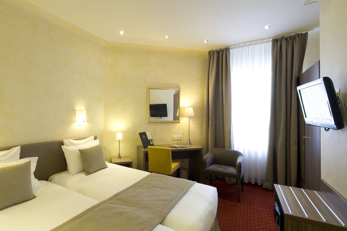 A hotel room with two beds, a desk, a chair, a TV, and a window with curtains.