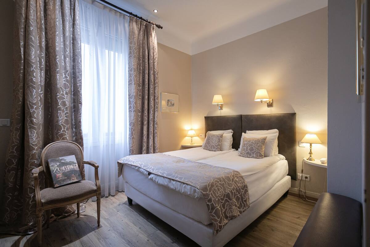 A hotel room with a large bed, two bedside lamps, a chair with a cushion, and a window with curtains.