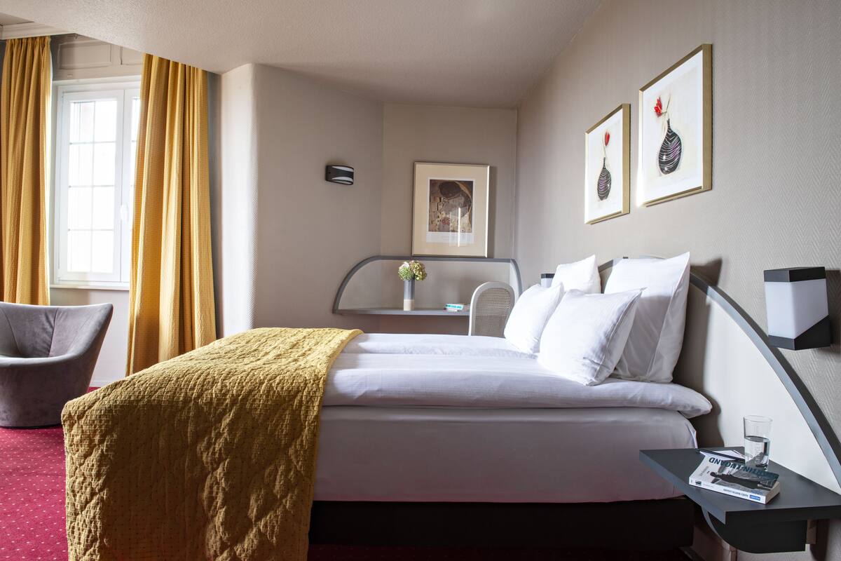 A hotel room with a bed, a chair, a bedside table with a magazine, and a window with curtains.