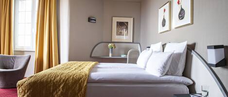 Une chambre d’hôtel avec un lit, une chaise, une table de chevet avec un magazine et une fenêtre avec des rideaux.