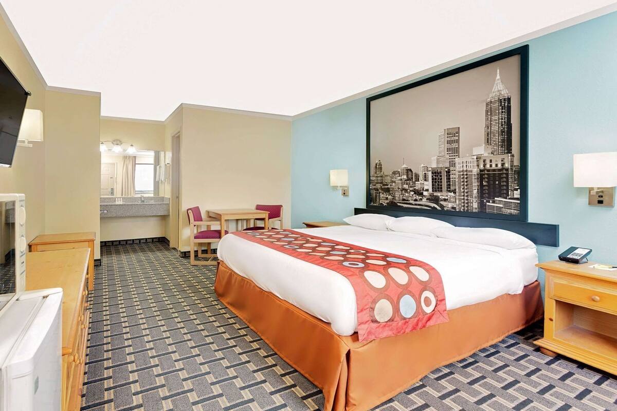 A hotel room with a bed, a desk, a chair, a television, and a cityscape painting on the wall.