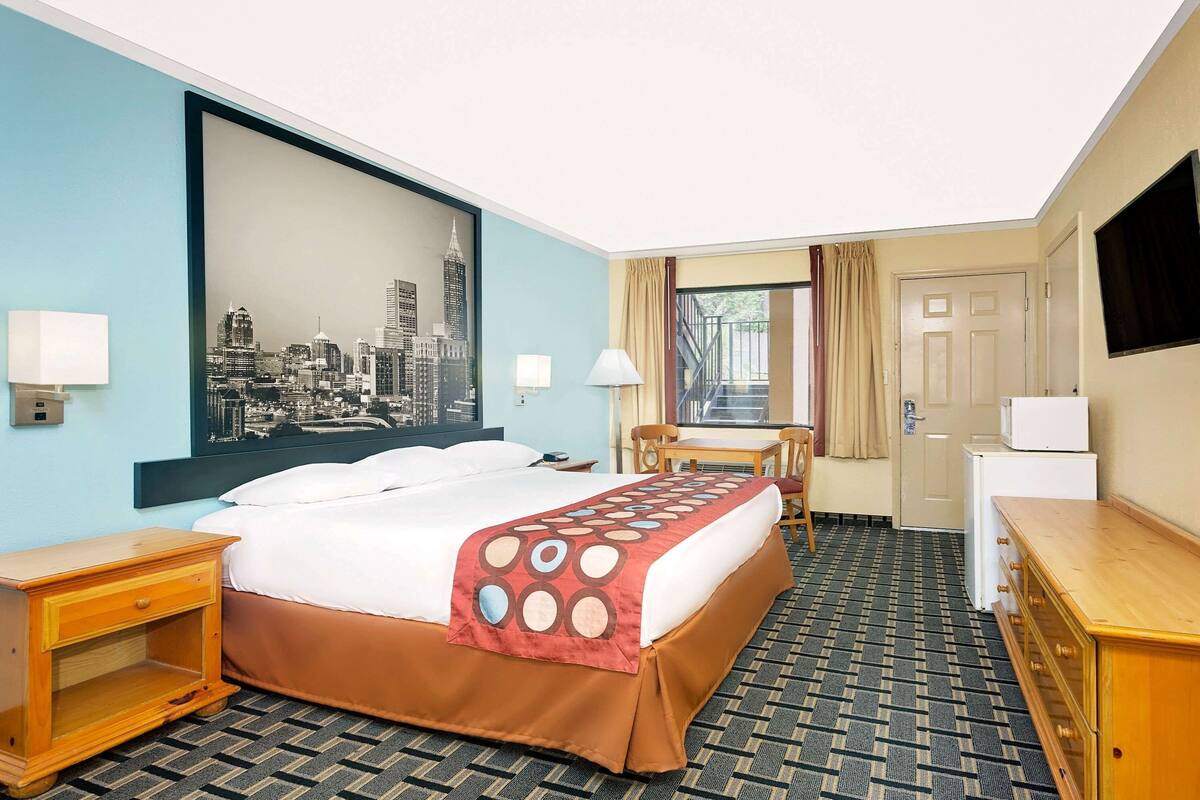 A hotel room with a bed, a desk, a chair, a TV, and a cityscape painting on the wall.