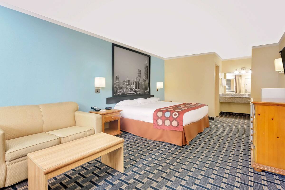 A hotel room with a bed, a sofa, a wooden bench, a nightstand, a TV, and a mirror.
