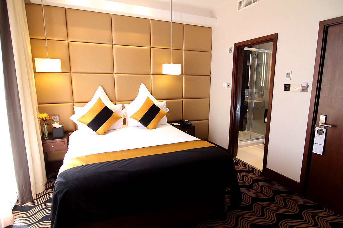 A hotel room with a large bed, a headboard, a bedside table with a lamp, and a door leading to a bathroom.