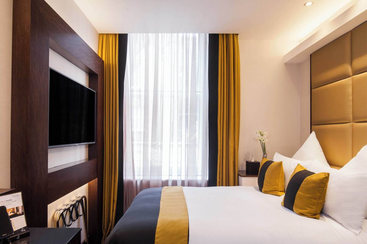 A hotel room with a large bed, a flat-screen TV mounted on the wall, a desk with a computer, and a window with curtains.