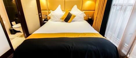 A hotel room with a large bed, a headboard with a light fixture, a nightstand, and a view of the outside through a window.
