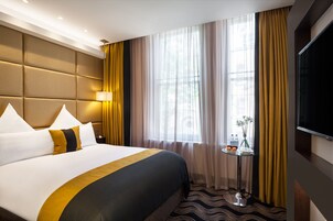 A hotel room with a large bed, a TV, a small table with a vase of flowers, and a window with curtains.