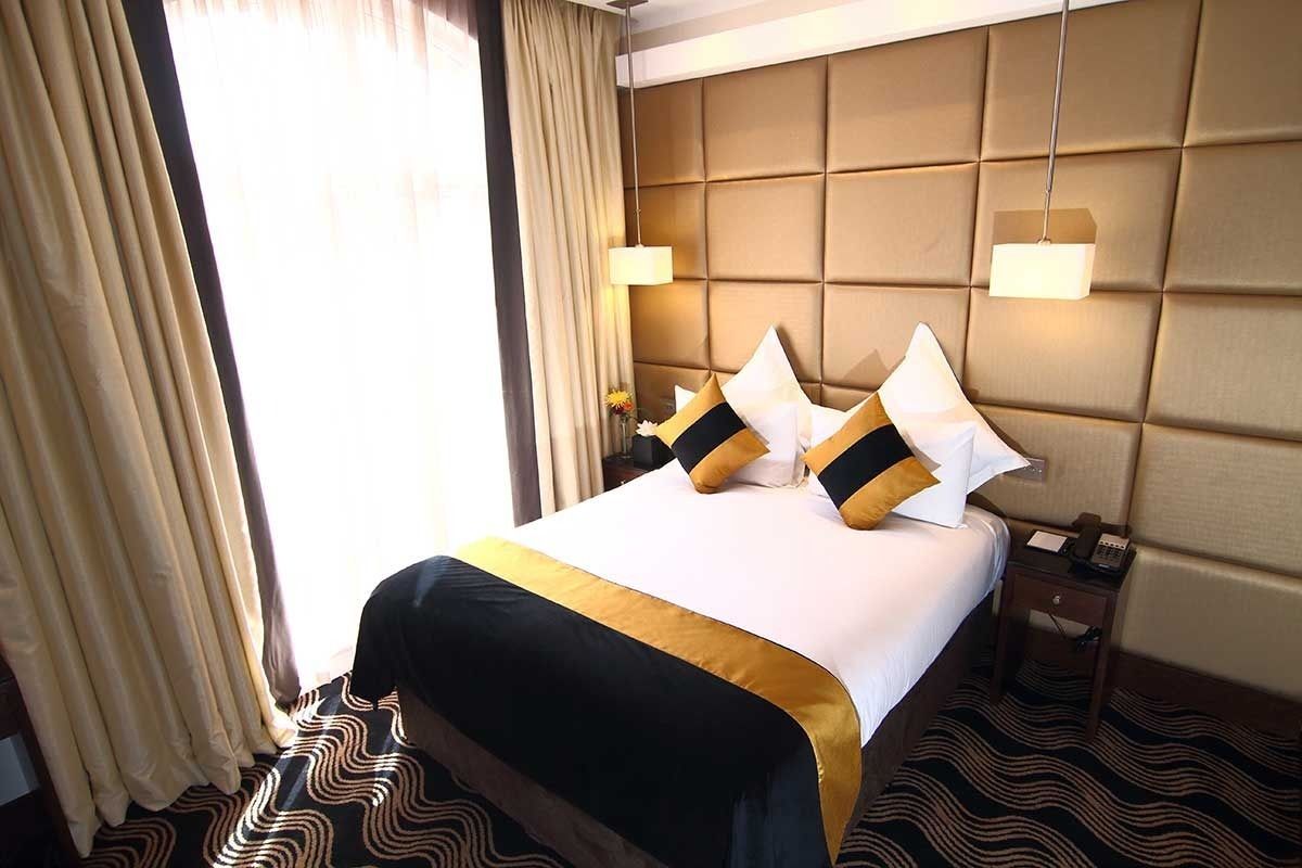 A hotel room with a large bed, a headboard with a padded design, two bedside lamps, a nightstand, and a patterned carpet.