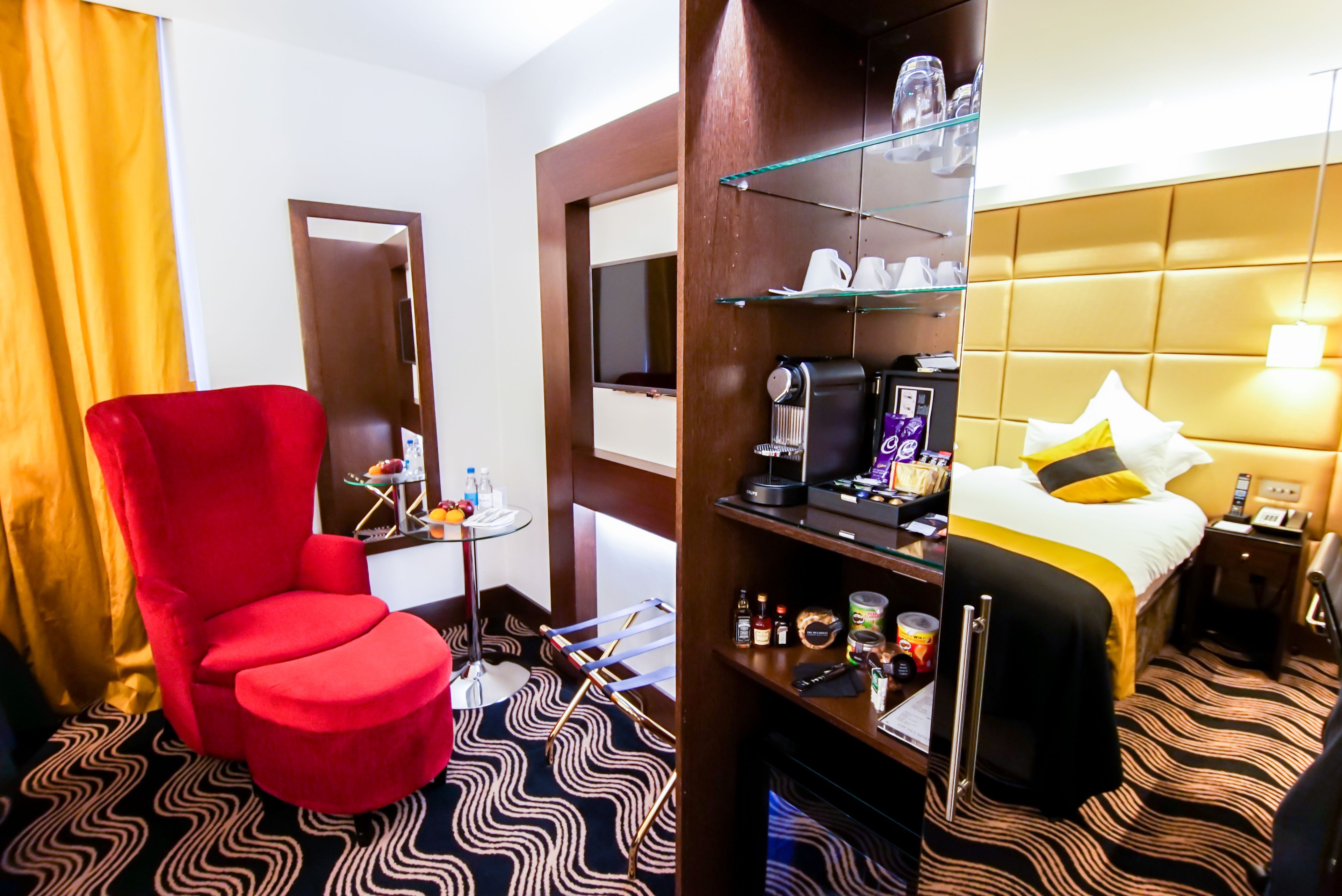 A hotel room with a red armchair, a bed with a yellow and black striped headboard, a minibar, a glass shelf with toiletries, and a patterned floor.