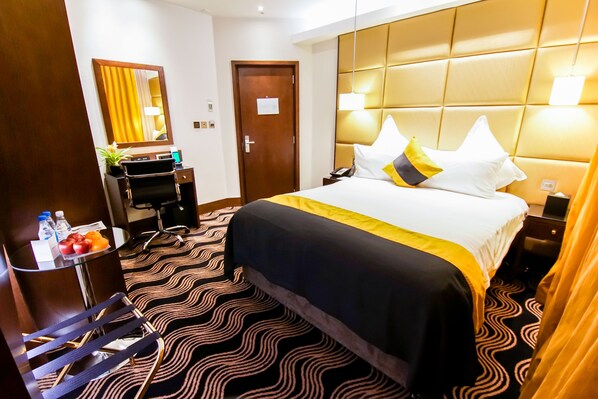 Habitación de hotel con una cama grande, un escritorio con espejo, una silla, una mesita con fruta y botellas, y una alfombra estampada.