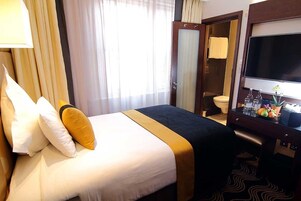 Habitación de hotel con cama, escritorio con televisor de pantalla plana, baño con inodoro y toalla colgada en la puerta, y ventana con cortinas.