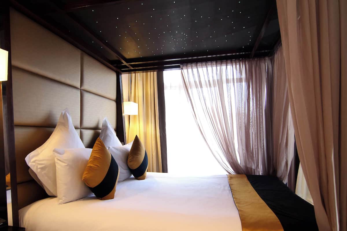 A hotel room with a bed, pillows, a nightstand, a lamp, and curtains.