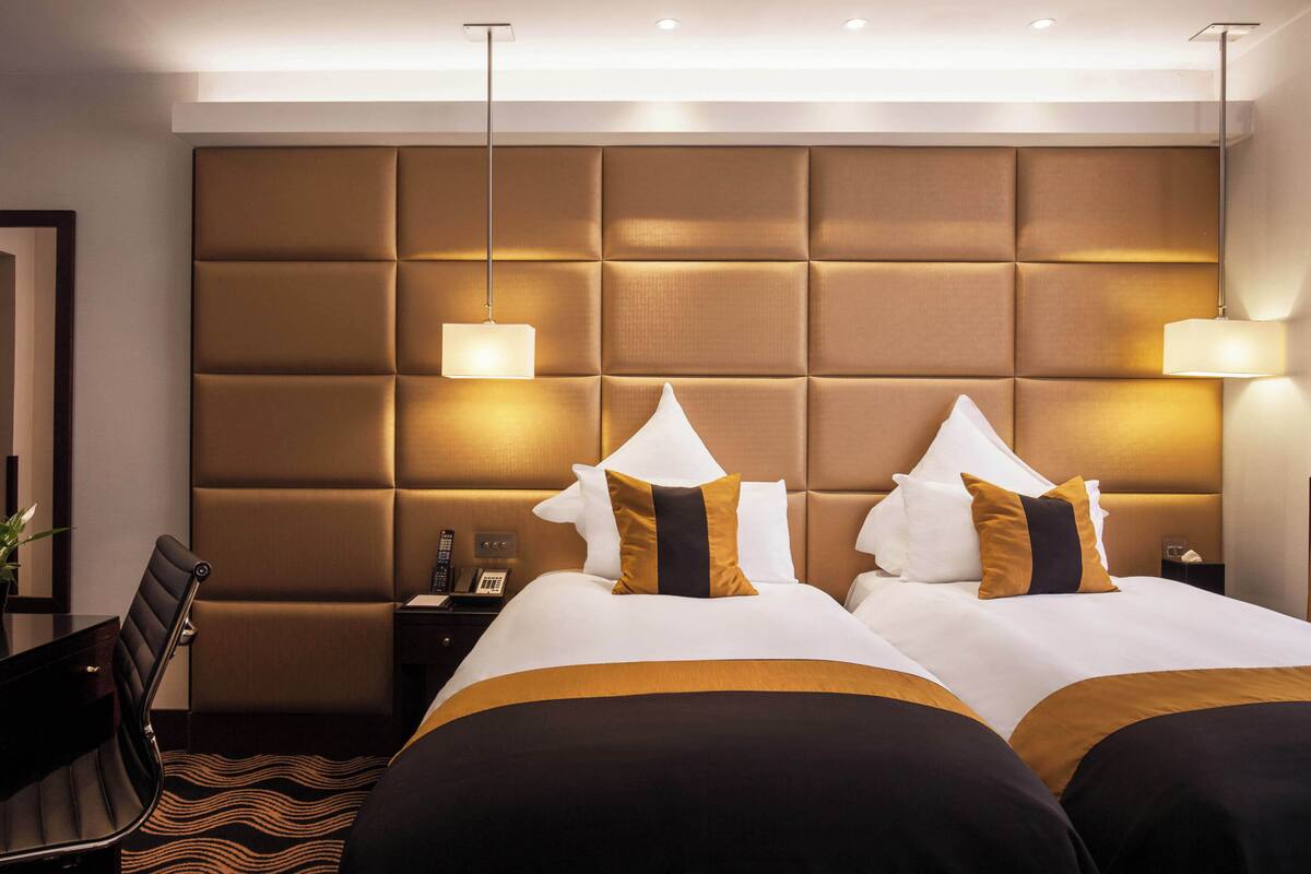 A hotel room with a large padded headboard, two beds with white and gold bedding, a desk with a chair, and bedside lamps.