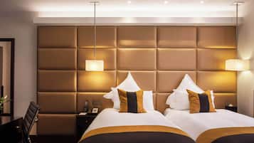 A hotel room with a large padded headboard, two beds with white and gold bedding, a desk with a chair, and bedside lamps.