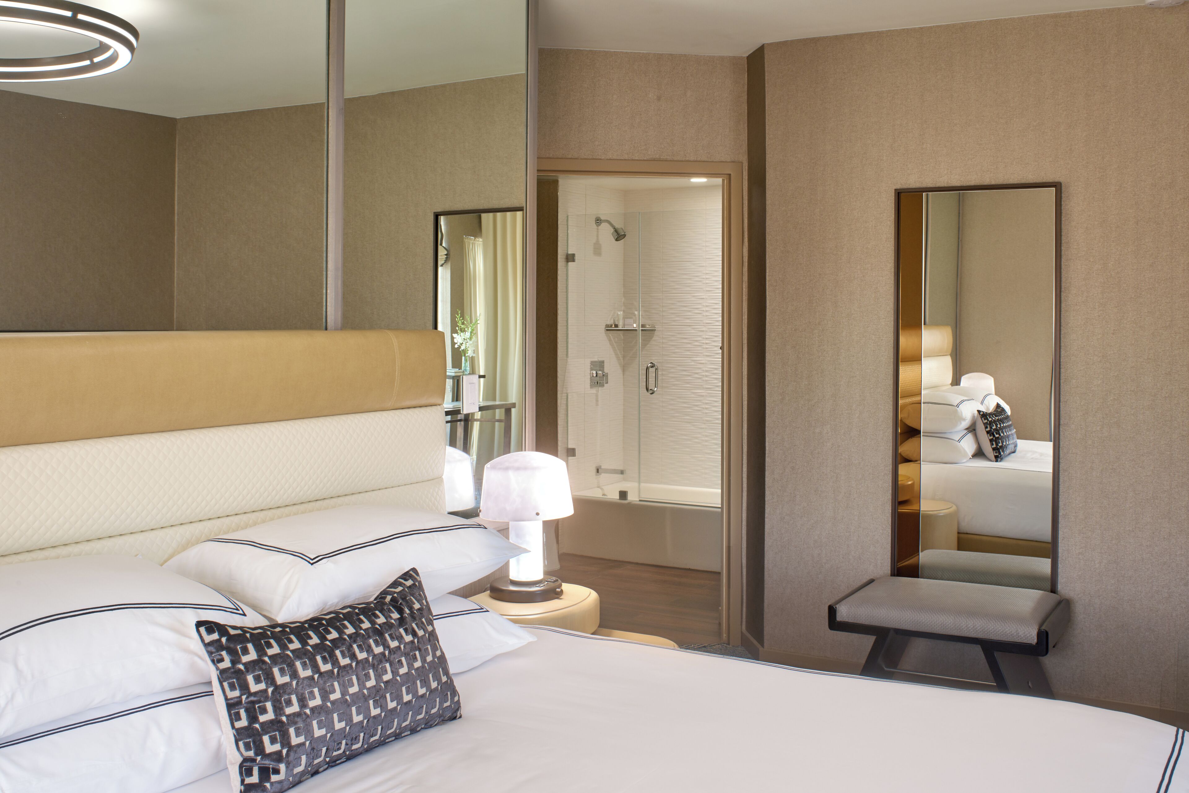 A hotel room with a bed, bedside lamp, a small table with a lamp, a bench, a shower area, and a large mirror.