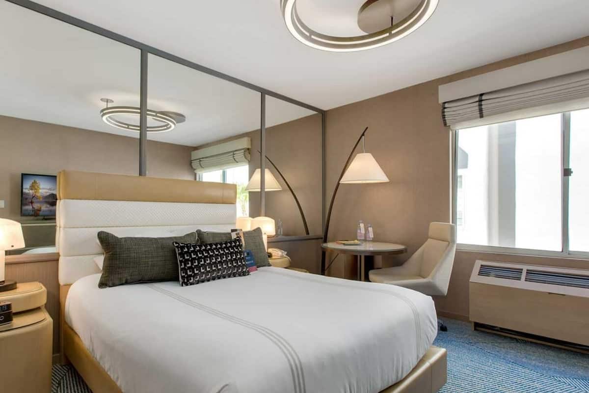 Une chambre d’hôtel avec un grand lit, des tables de chevet, une chaise, une petite table, une lampe, une fenêtre et un luminaire au plafond.