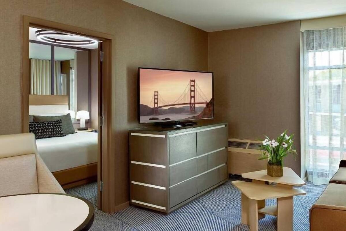 A hotel room with a bed, a TV displaying the Golden Gate Bridge, a desk, a chair, and a window with curtains.