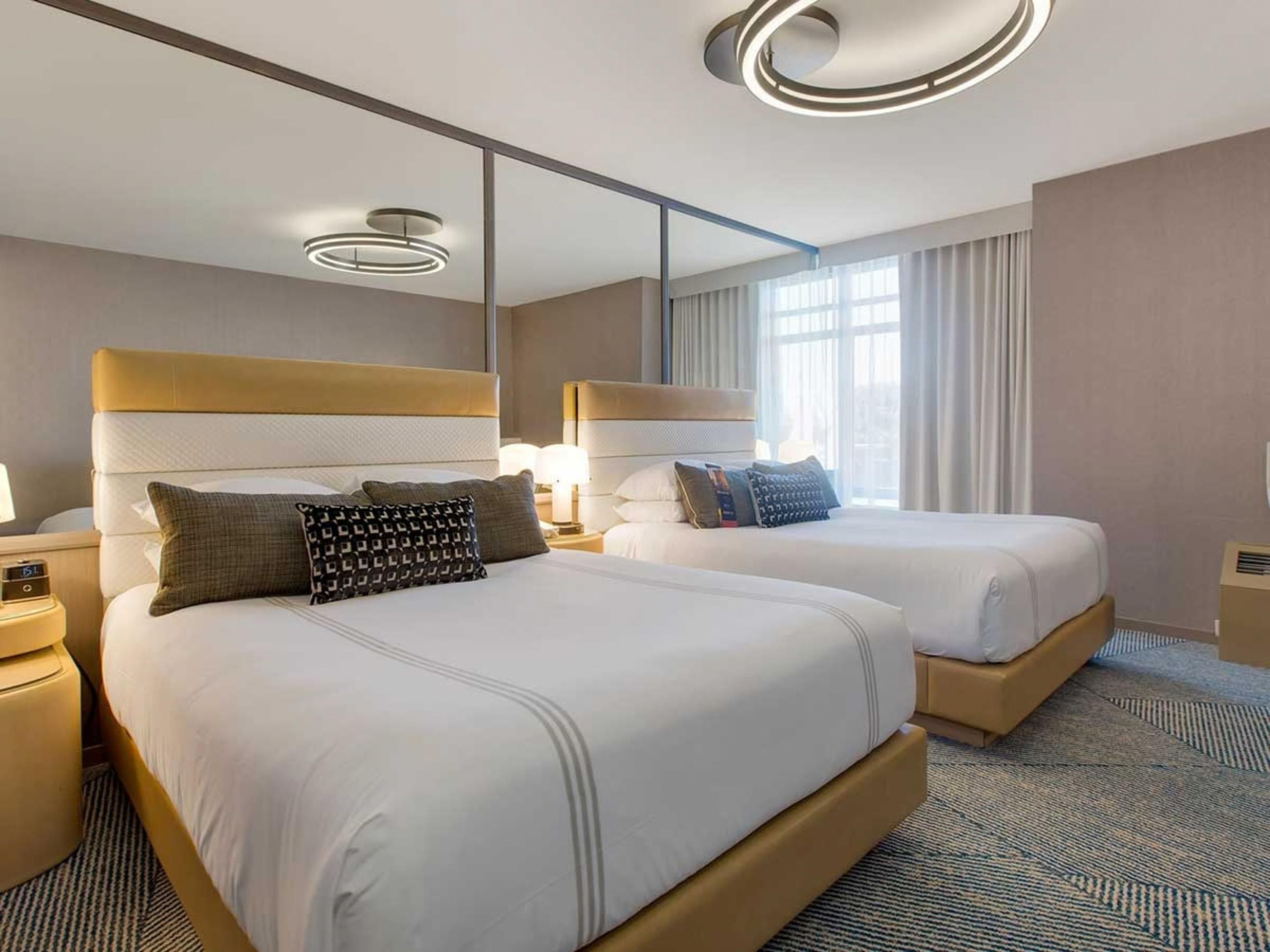 A hotel room with two beds, a large mirror, and a modern ceiling light fixture.