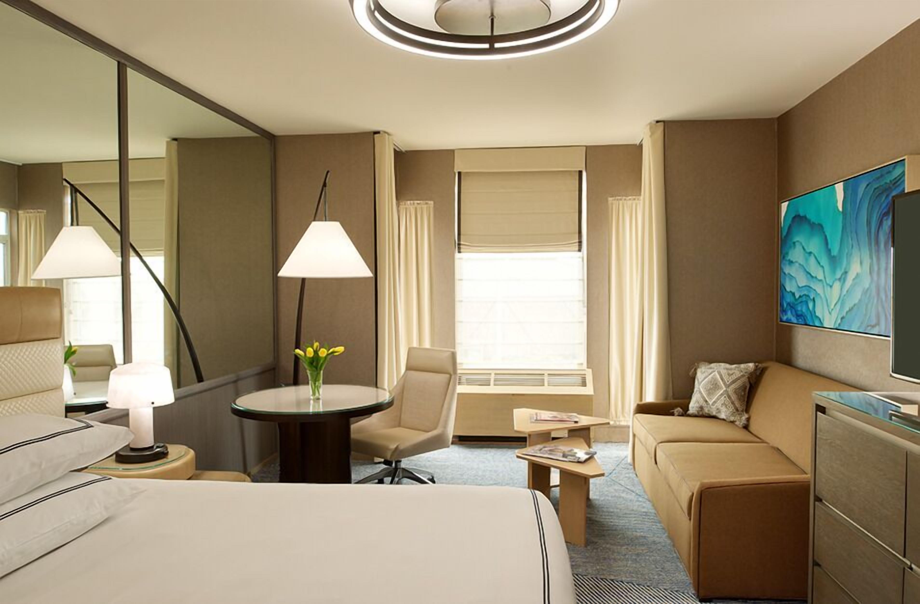 Ein Hotelzimmer mit einem Bett, einer Couch, einem kleinen Tisch, einem Sessel, einem Fernseher und einem Fenster mit Vorhängen.