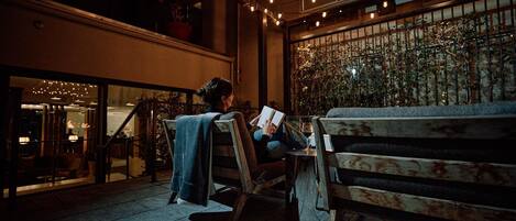 A person sitting on a wooden bench reading a book in an outdoor area with string lights and plants.