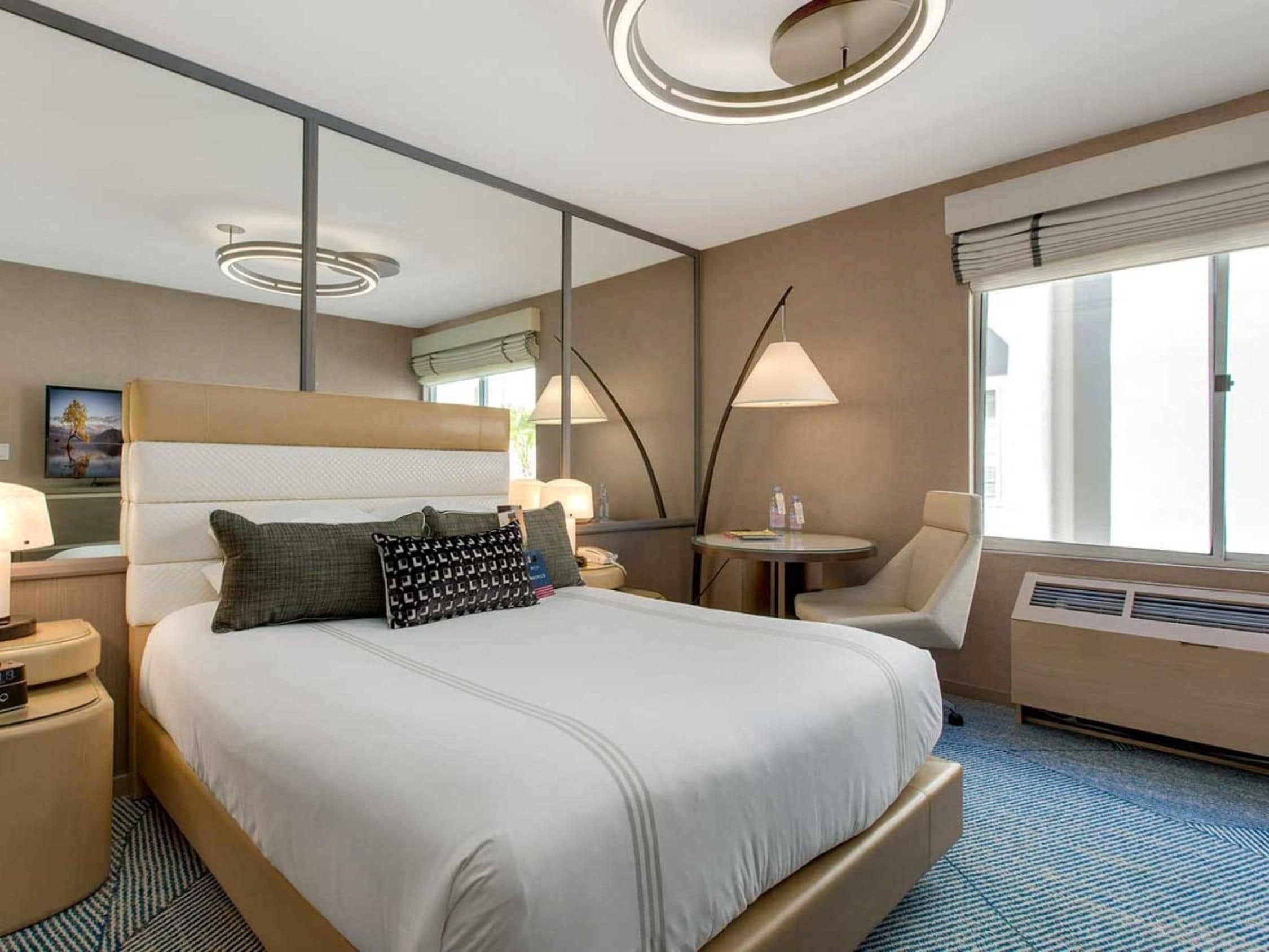 Una habitación de hotel con una cama grande, una mesita de noche con lámpara, una silla, una pequeña mesa con bebidas y una ventana con persianas.