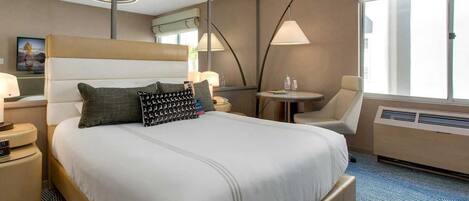 Una habitación de hotel con una cama grande, una mesita de noche con lámpara, una silla, una pequeña mesa con bebidas y una ventana con persianas.