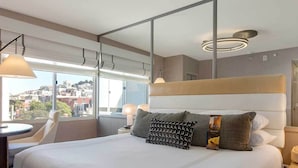 A hotel room with a large bed, a desk, and a chair. There is a view of the city through the window.
