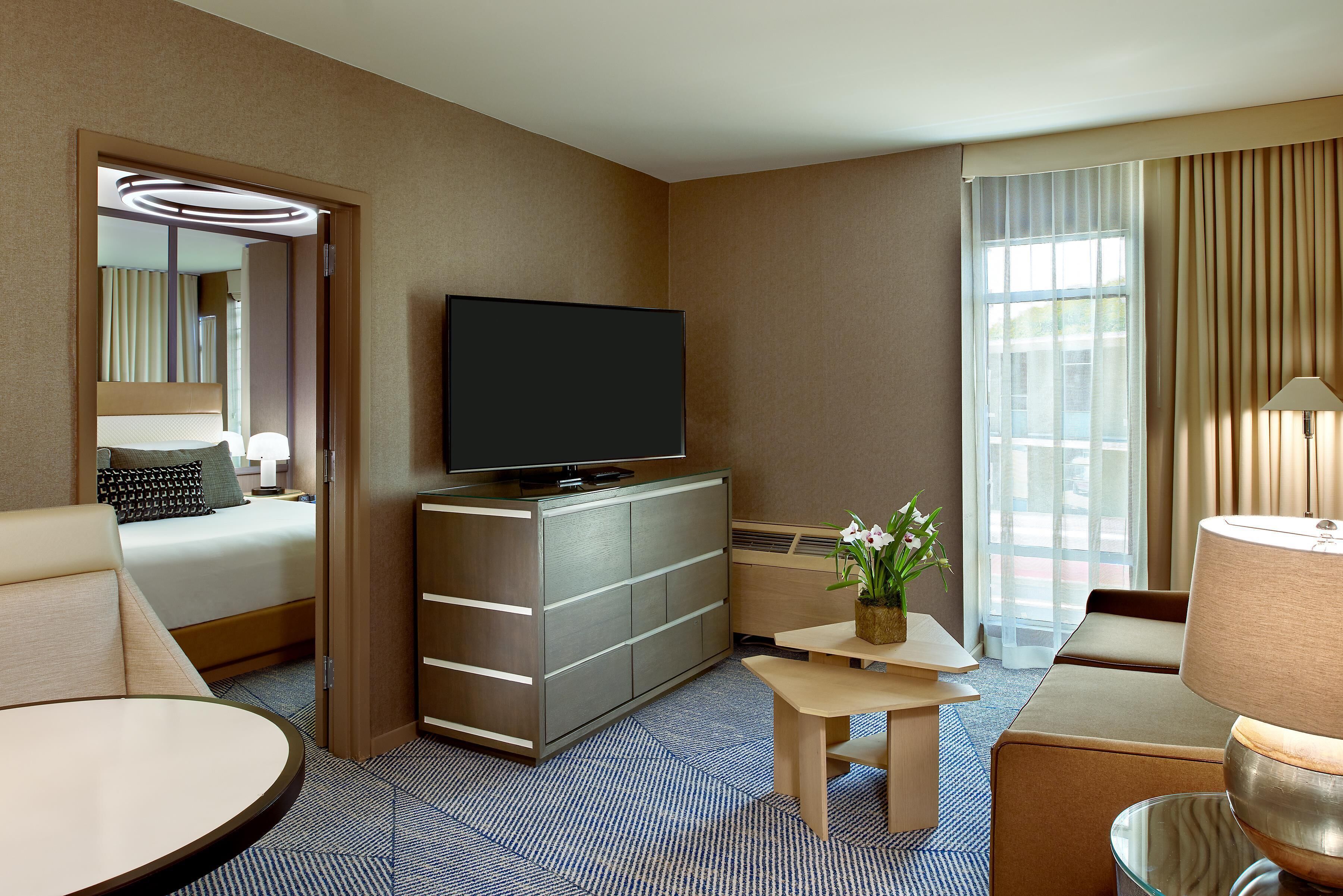 A modern hotel room with a flat-screen TV on a cabinet, a bed with a headboard, a small table with a vase of flowers, and a large window with curtains.