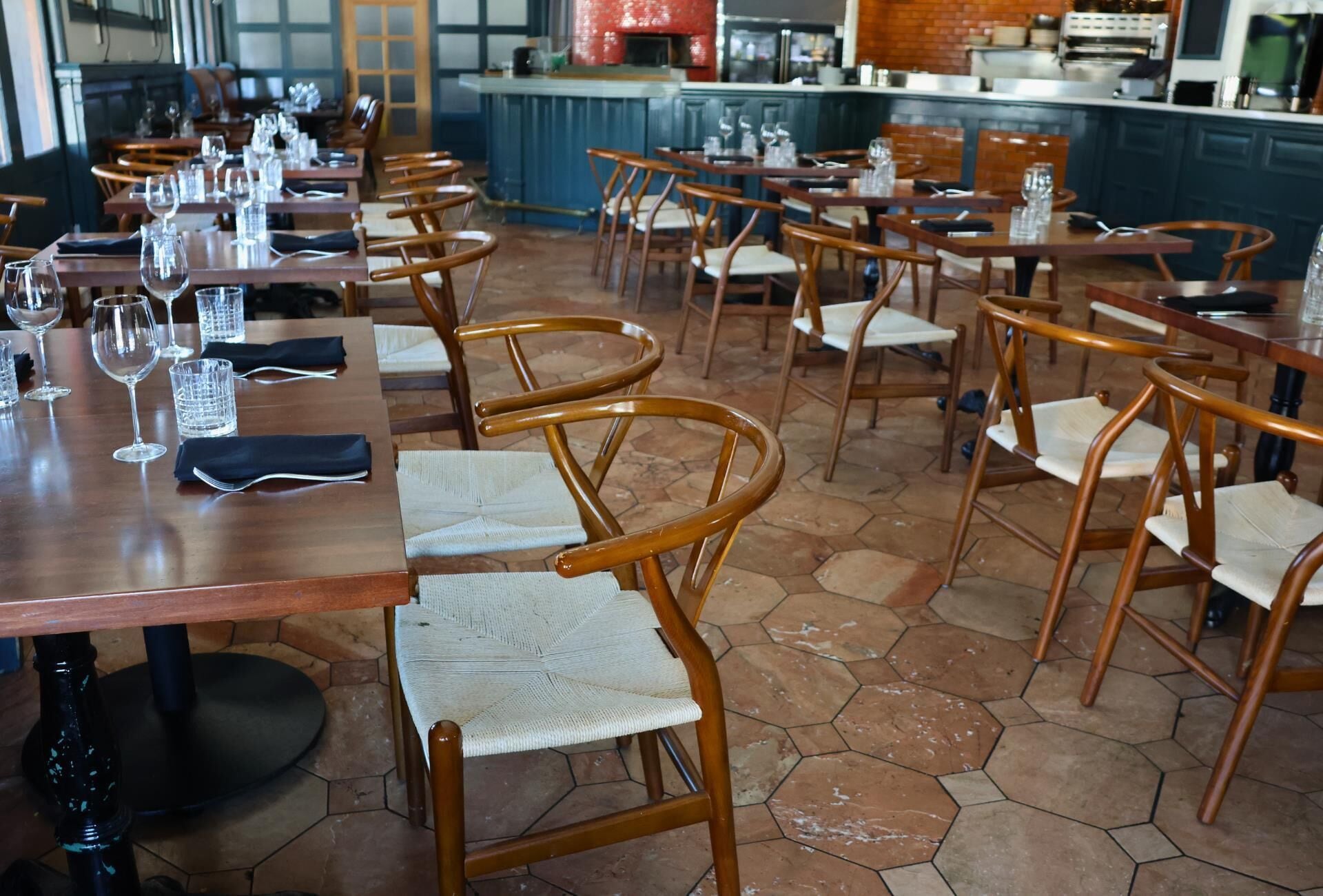 Un restaurante con mesas y sillas de madera, suelo de baldosas y una cocina al fondo.