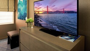 A hotel room with a large flat-screen TV on a wooden cabinet, a small stool, and a framed abstract painting on the wall.