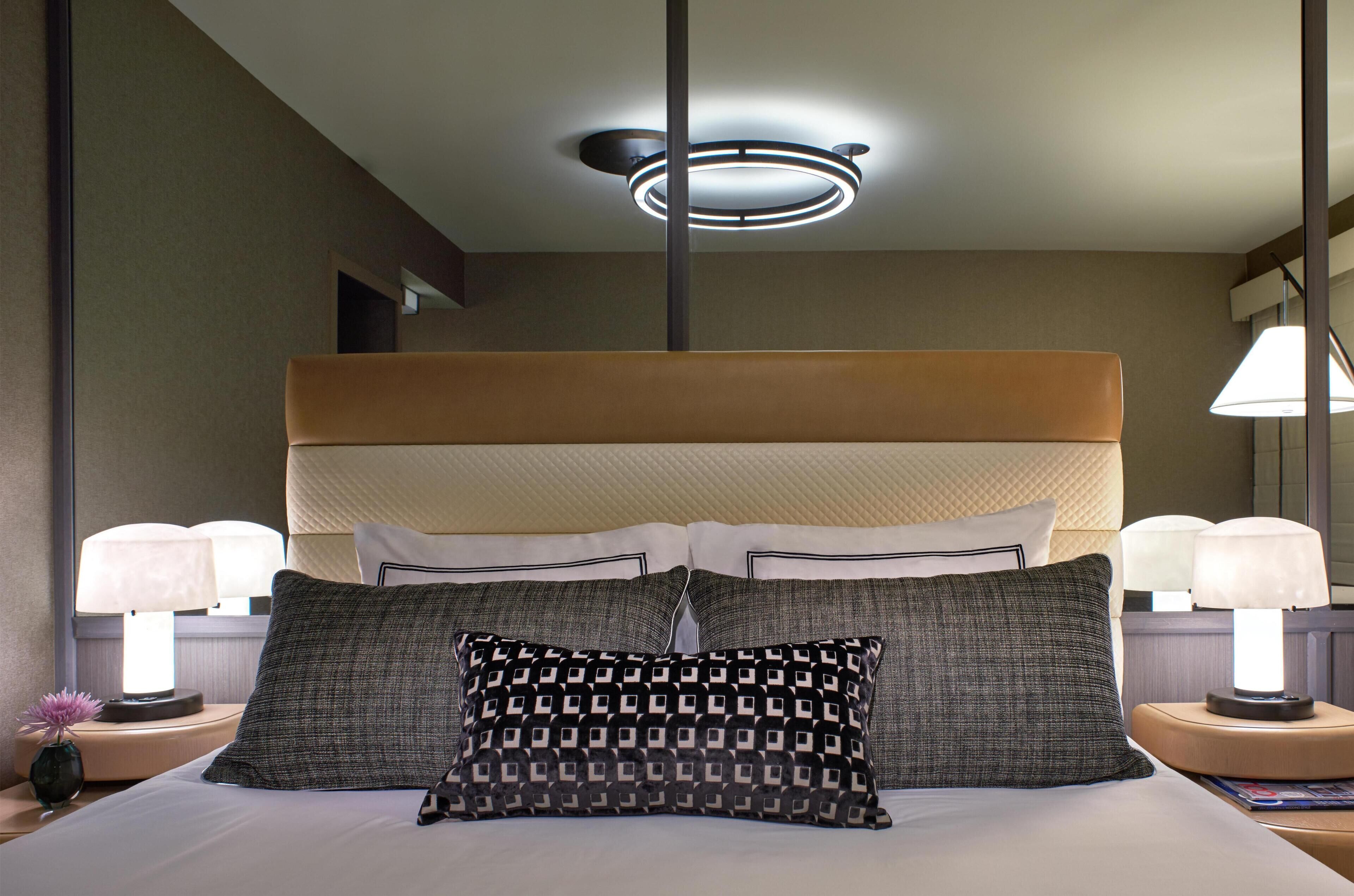 Habitación de hotel con una cama grande, dos lámparas de noche, una lámpara de techo moderna y una mesita de noche con un jarrón y una lámpara.