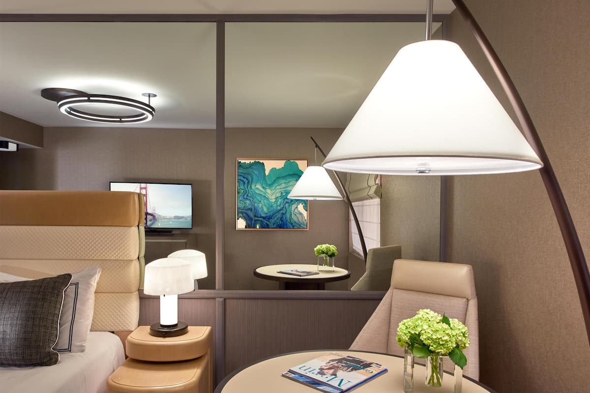 Une chambre d’hôtel moderne avec un lit, une table de chevet avec une lampe, une table ronde avec un magazine et des fleurs, un tableau au mur et un téléviseur.