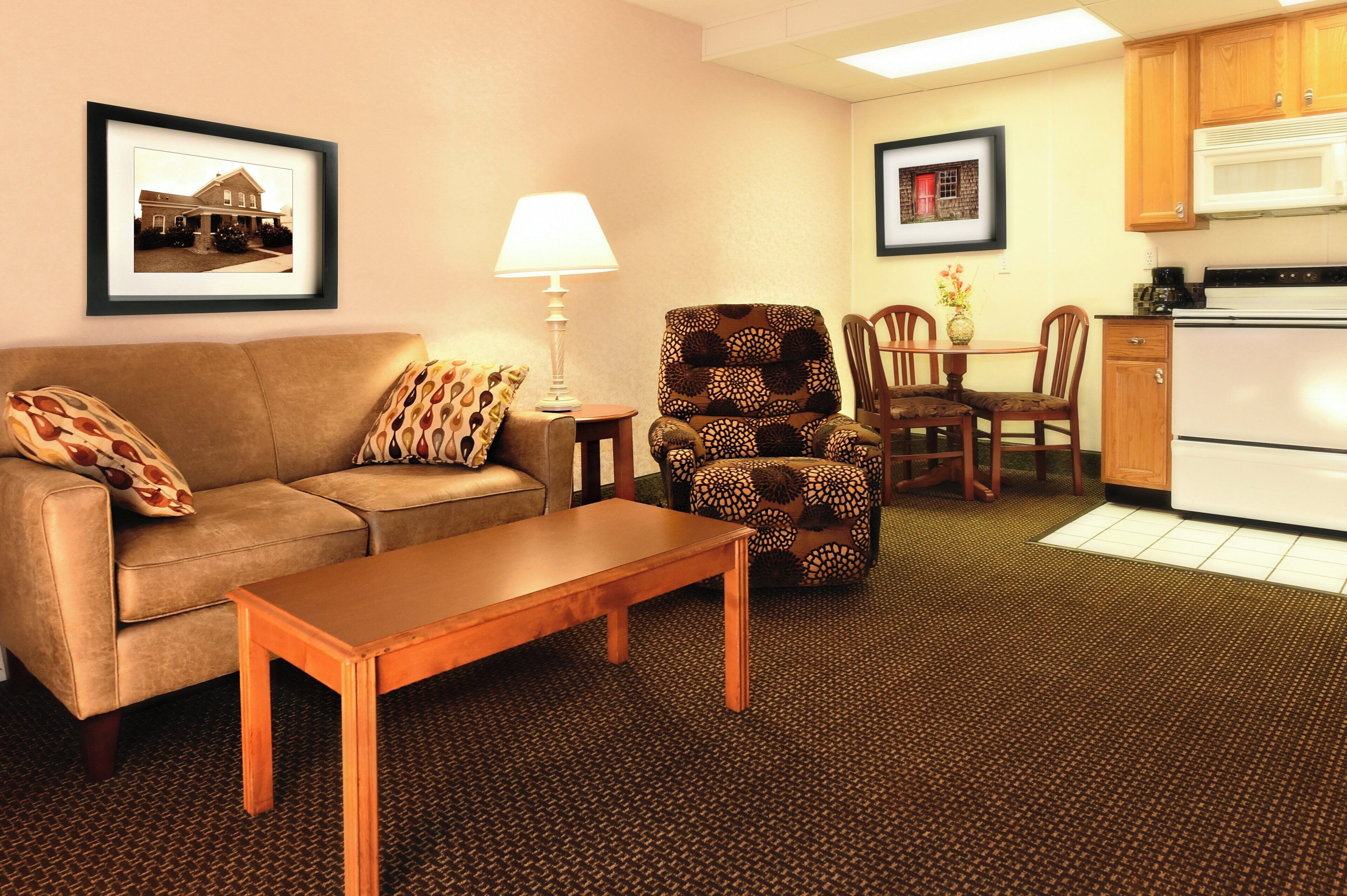 A living room with a sofa, a patterned armchair, a wooden coffee table, a lamp, a dining table with chairs, and a kitchen area with a microwave and stove.