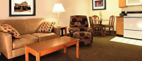 A living room with a sofa, a patterned armchair, a wooden coffee table, a lamp, a dining table with chairs, and a kitchen area with a microwave and stove.